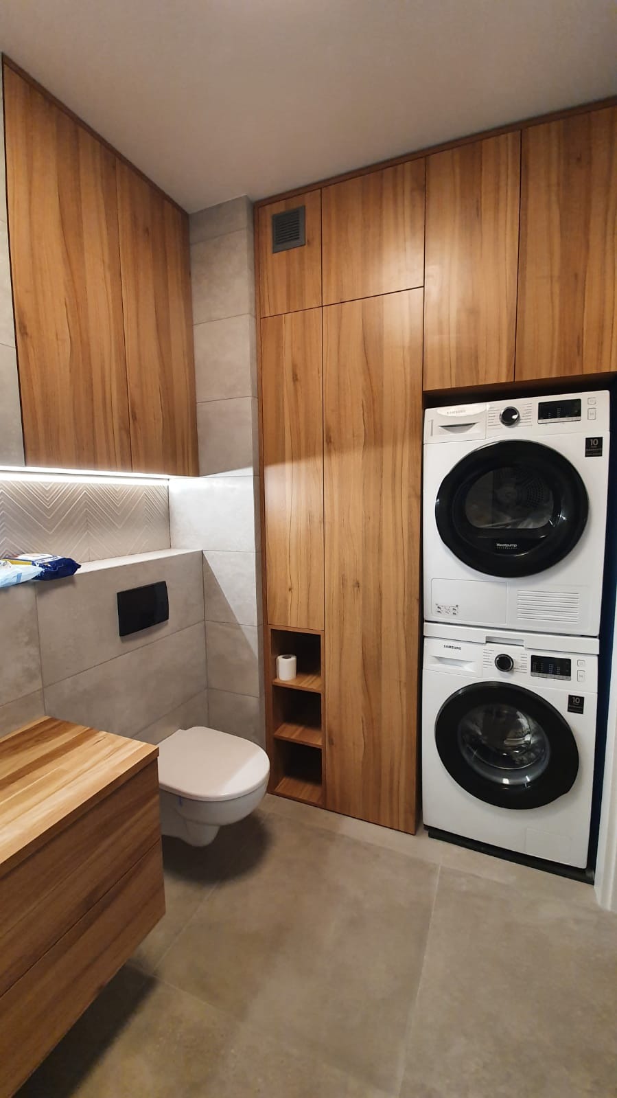 Nowoczesna łazienka z drewnianymi szafkami, zabudowaną pralką i suszarką Samsung, oraz toaletą w minimalistycznym stylu.