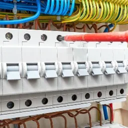 Ręka przykręca śrubkę w rozdzielnicy elektrycznej z bezpiecznikami za pomocą czerwono-czarnego śrubokręta, widoczne kolorowe przewody.