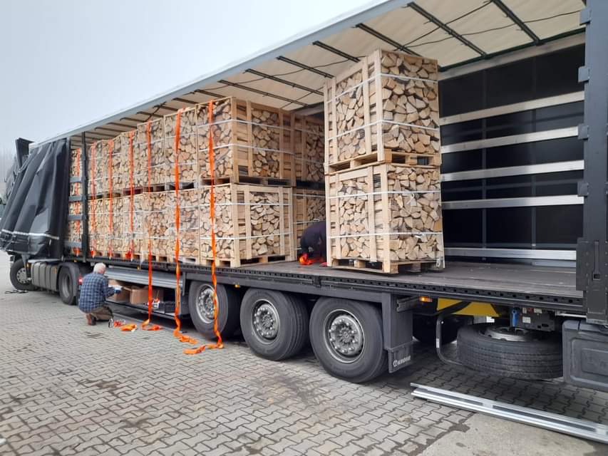 Załadunek pociętego drewna w skrzyniopaletach na naczepę ciężarówki, zabezpieczonego pomarańczowymi pasami transportowymi.