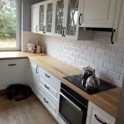 Biała kuchnia w stylu klasycznym z drewnianym blatem, białymi kaflami i śpiącym psem rasy owczarek niemiecki przy dolnych szafkach