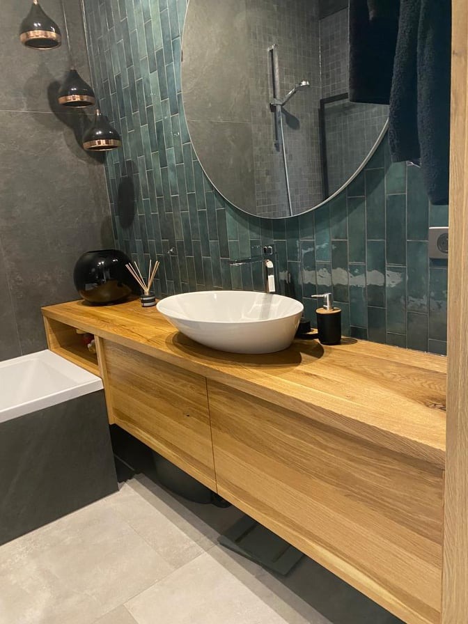 Nowoczesna łazienka z drewnianą szafką pod umywalką nablatową, okrągłym lustrem i turkusowymi płytkami na ścianie.