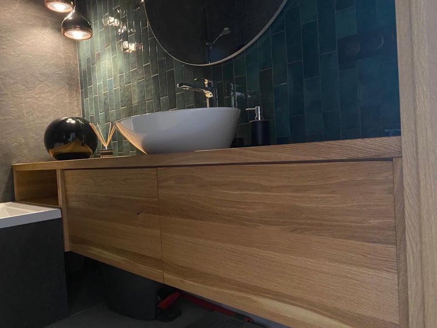 Nowoczesna łazienka z drewnianą szafką pod umywalką nablatową, zielone płytki na ścianie i okrągłe lustro z oświetleniem.