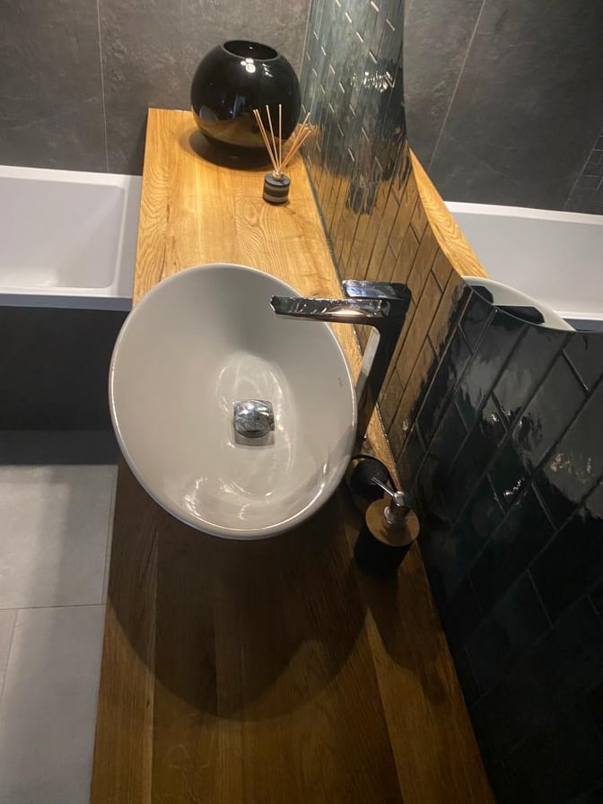 Nowoczesna łazienka z umywalką nablatową na drewnianym blacie, czarne płytki na ścianie, minimalistyczne dodatki: czarny wazon i dozownik mydła.