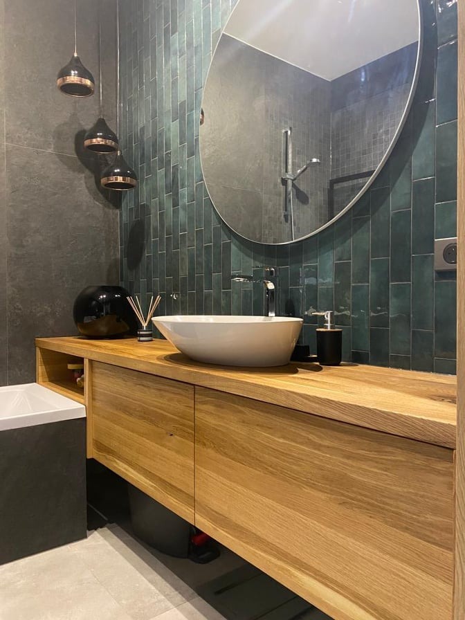 Nowoczesna łazienka z drewnianą szafką pod umywalką, okrągłym lustrem i zielonymi płytkami na ścianie, oświetlona trzema wiszącymi lampami z miedzianymi detalami.