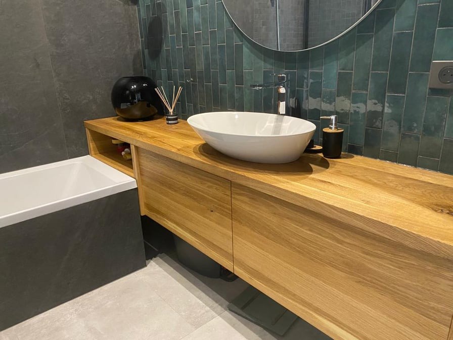 Nowoczesna łazienka z drewnianą szafką pod umywalkę nablatową, zielone płytki na ścianie i czarna wanna obok.