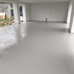 MAREXLUX - Świeżo wylana, lśniąca, jasnoszara wylewka samopoziomująca w pustym pomieszczeniu z białymi ścianami i widocznym placem budowy na zewnątrz.
