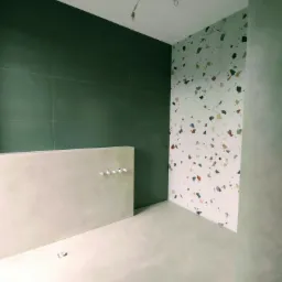 Wnętrze łazienki w trakcie wykańczania: zielona ściana z płytek, ściana z wzorem lastryko, surowa wylewka na podłodze, widoczne punkty świetlne i wystające kable.