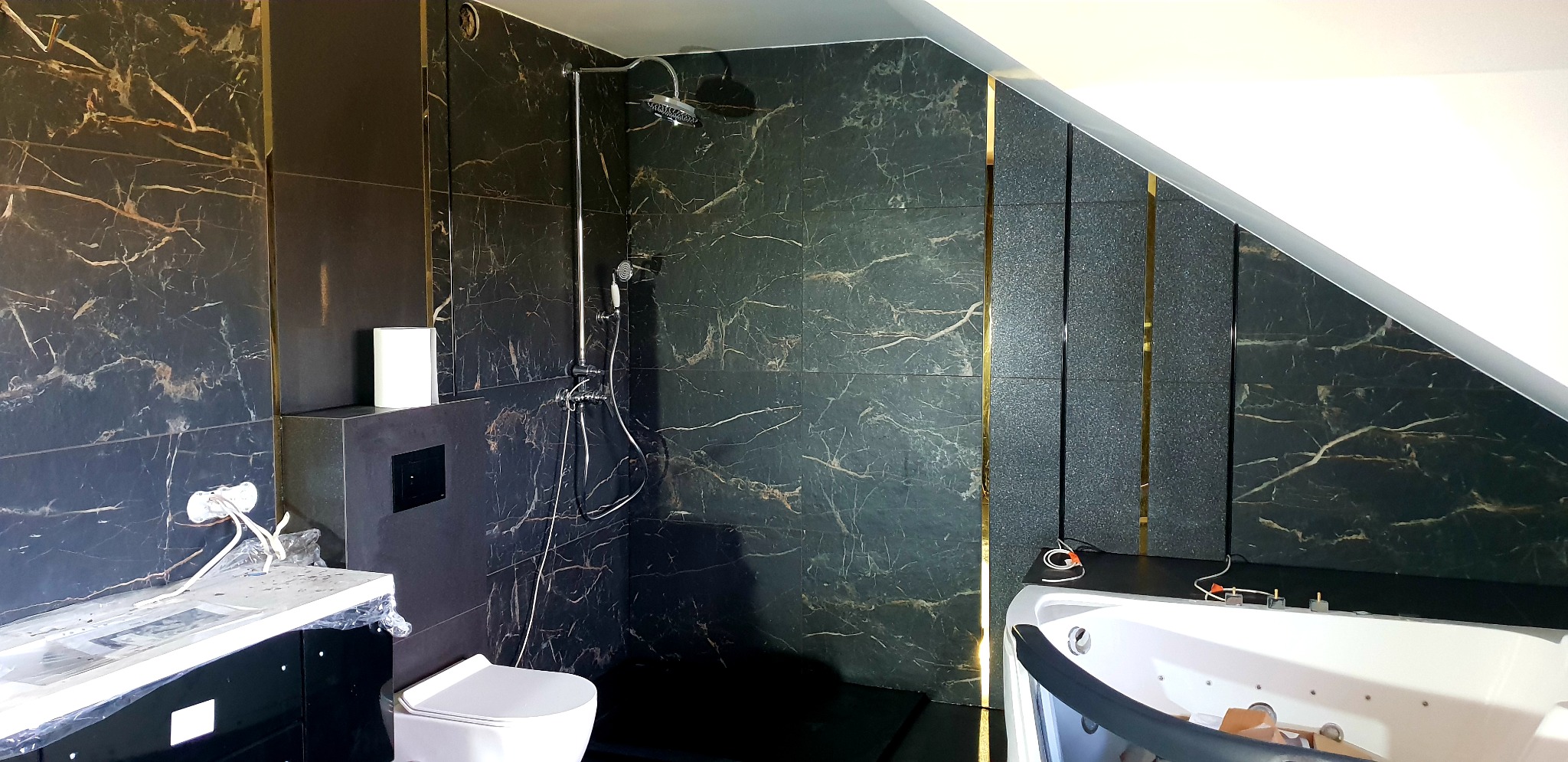 Nowoczesna łazienka na poddaszu z czarnymi płytkami imitującymi marmur, złotymi akcentami, białą armaturą i wanną z hydromasażem w trakcie prac wykończeniowych, widoczne wystające przewody.