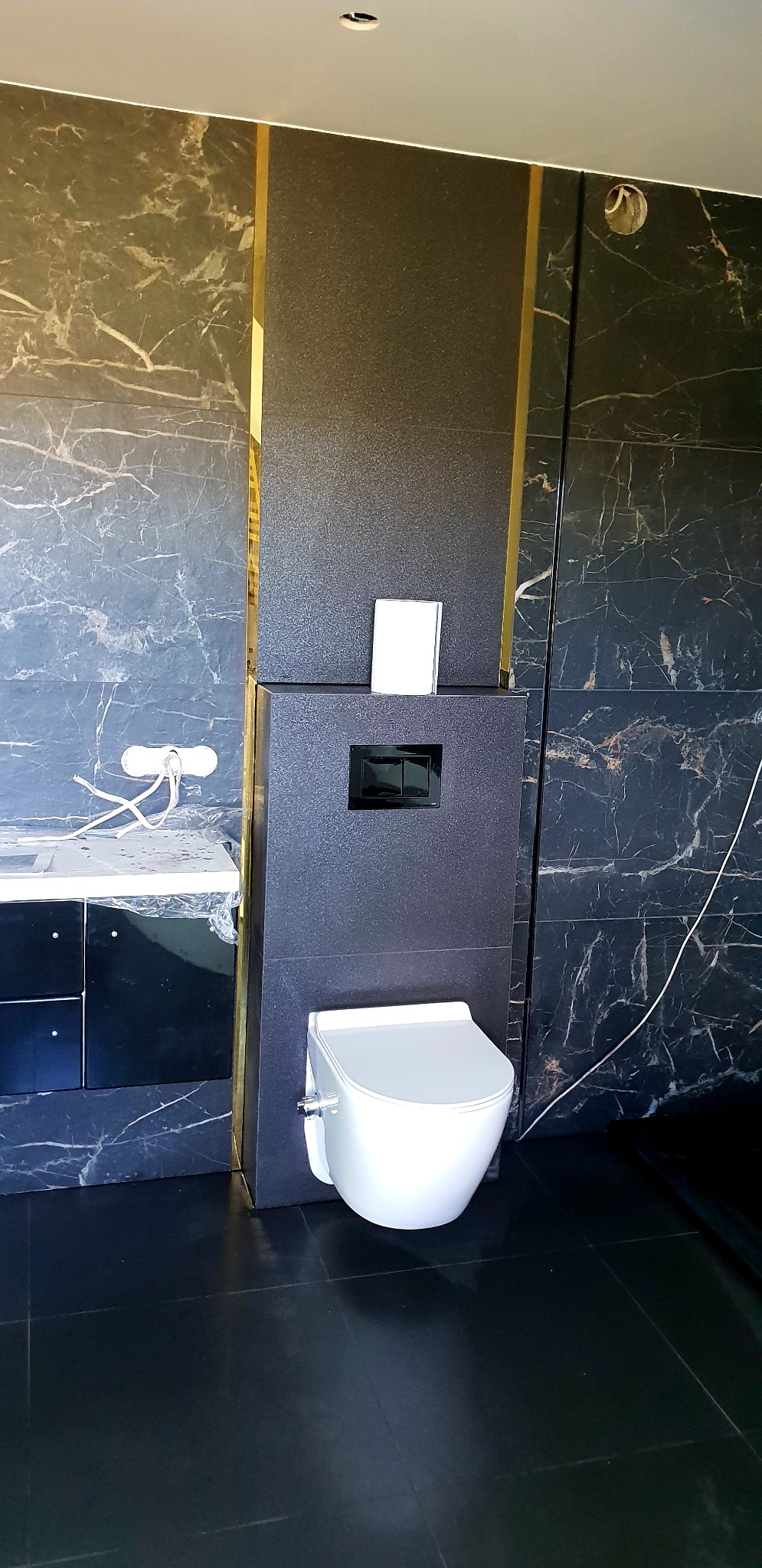 Elegancka, nowocześnie wykończona łazienka z czarnymi płytkami imitującymi marmur z białymi żyłami, złotymi akcentami i podwieszaną toaletą z czarnym przyciskiem spłukiwania.