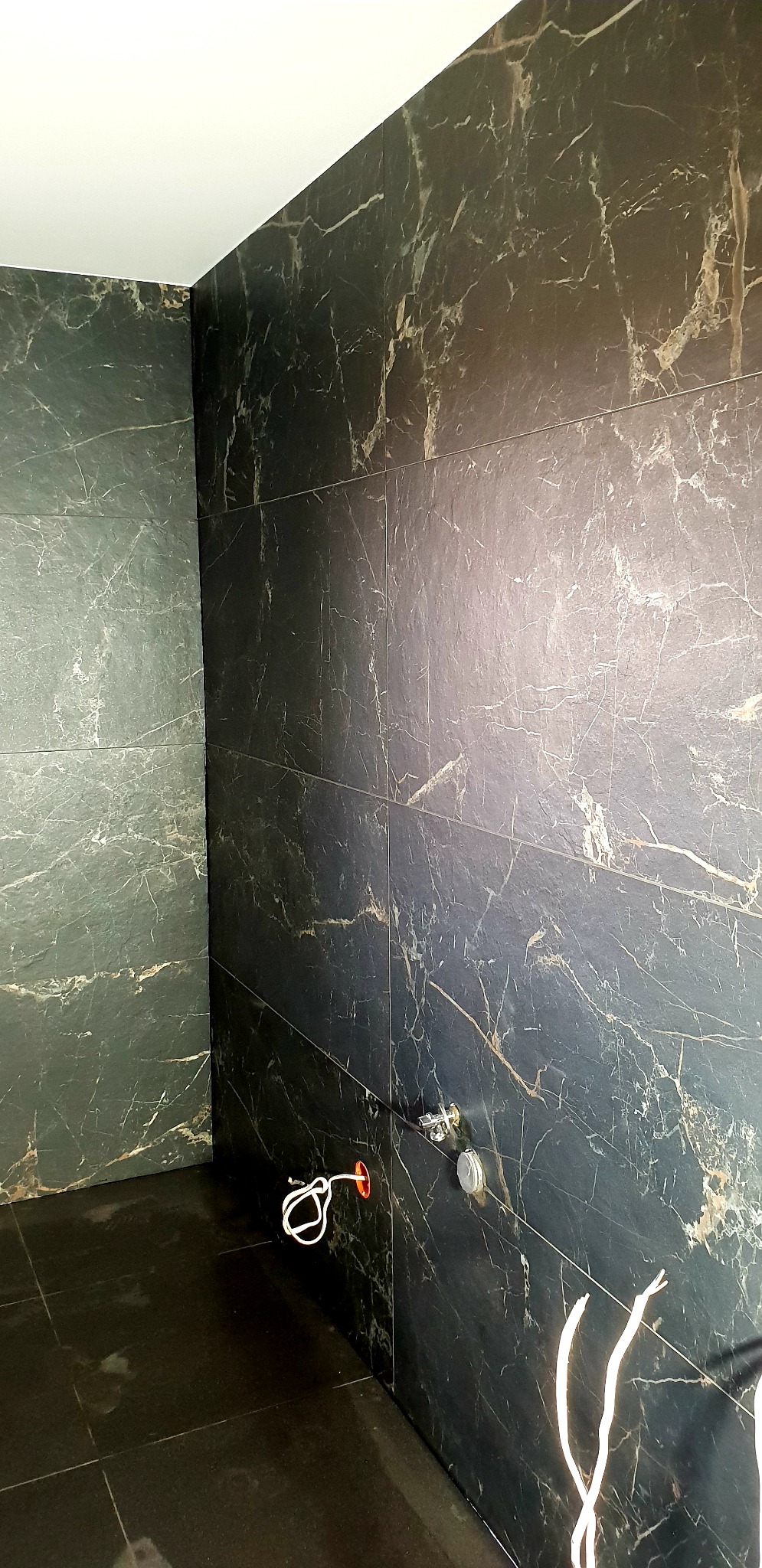 Łazienka w trakcie remontu: ściany i podłoga wyłożone ciemnymi płytkami imitującymi marmur z jasnymi żyłami, widoczne wystające kable i przyłącze wodne.