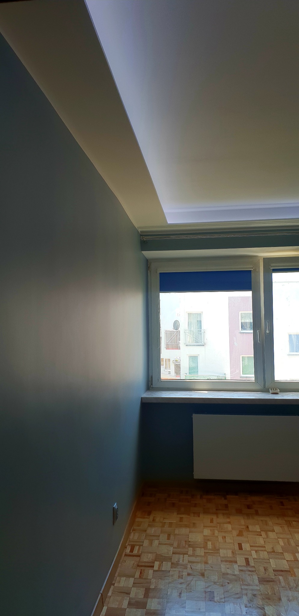 Świeżo pomalowane na szaro wnętrze pokoju z parkietem, widok na okno z roletą i zabudowaną listwą sufitową z oświetleniem LED.