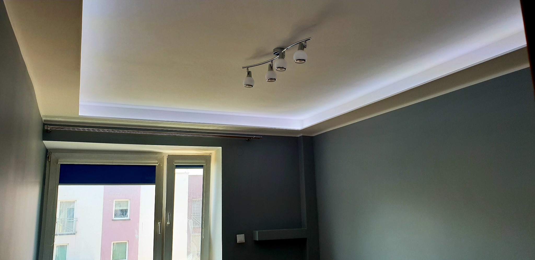 Pokój z szarą ścianą, oświetleniem LED wokół podwieszanego sufitu i lampą sufitową z czterema reflektorami, widok na okno z roletą.