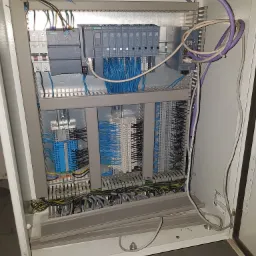 Otwarta szafa sterownicza z modułami Siemens, licznymi przewodami niebieskimi i fioletowymi, widoczne złączki i bezpieczniki, szyna DIN.