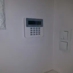 Biały panel sterowania alarmu domowego Satel Perfecta zamontowany na ścianie, obok włącznik światła, widoczny fragment dekoracyjnej listwy sufitowej i kąt ściany przy podłodze z drobnymi...