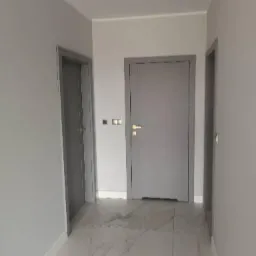 Wnętrze korytarza z dwiema parami drzwi w szarych ościeżnicach, marmurowa podłoga z szarymi żyłami, ściany pomalowane na jasny odcień szarości.