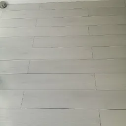 Jasne, matowe panele podłogowe ułożone wzdłuż ściany z białą listwą przypodłogową, widoczny metalowy odbojnik.