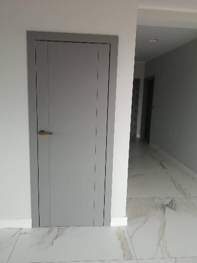 Zamknięte, szare drzwi wewnętrzne z minimalistyczną, złotą klamką, osadzone w białej ścianie, z widocznym fragmentem marmurowej podłogi i perspektywą korytarza.