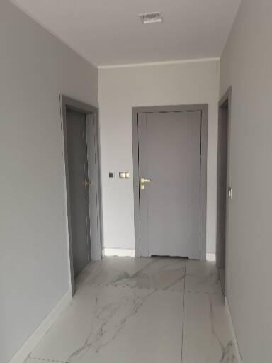 Wnętrze korytarza z dwiema parami drzwi w szarych ościeżnicach, marmurowa podłoga z szarymi żyłami, ściany pomalowane na jasny odcień szarości.