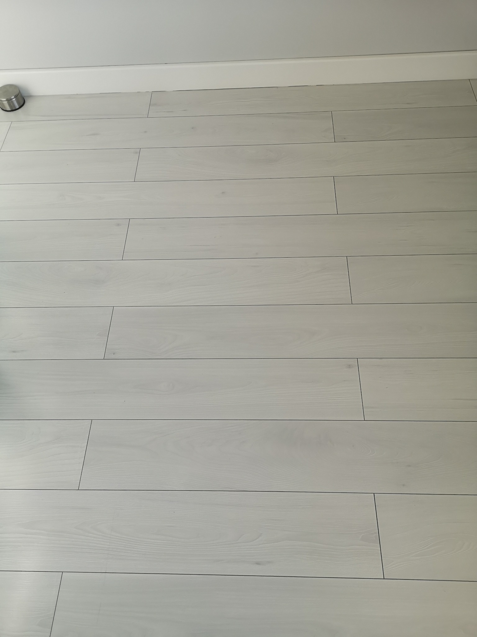 Jasne, matowe panele podłogowe ułożone wzdłuż ściany z białą listwą przypodłogową, widoczny metalowy odbojnik.