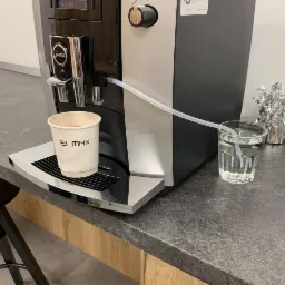 Ekspres do kawy Jura E6 podłączony do wody z zewnątrz za pomocą przezroczystego wężyka, filiżanka kawy na podstawce, w tle szklanka wody i dekoracja.