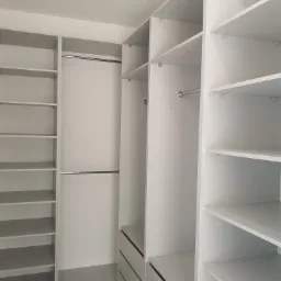 Wnętrze garderoby z białymi półkami i szufladami, widoczne drążki na ubrania, minimalistyczny design.