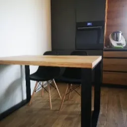 Stół z blatem z jasnego drewna i czarnymi nogami, dwie czarne krzesła z drewnianymi nogami, w tle zabudowa kuchenna w ciemnych kolorach.