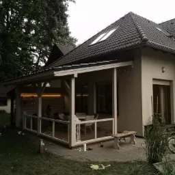 Zadaszony taras przy domu z drewnianą konstrukcją, szklanymi panelami i oświetleniem LED, widoczne narzędzia i materiały sugerujące prace wykończeniowe.