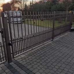 Otwarta, metalowa, brązowa brama wjazdowa z automatyką na posesję wyłożoną kostką brukową, w tle widoczne drzewa i dom jednorodzinny.