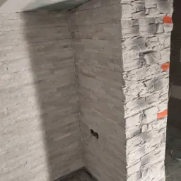 Wykończony narożnik ściany z dekoracyjną, jasną okładziną imitującą kamień, widoczne niedoskonałości na łączeniu z sufitem i wystające kable elektryczne.