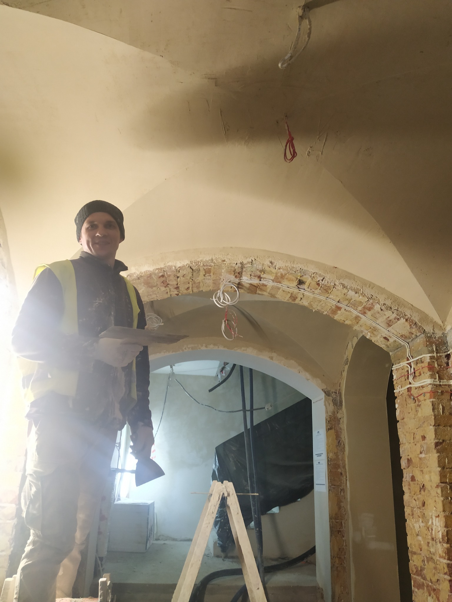 Mężczyzna w kamizelce odblaskowej, stojący na drabinie, trzymający szpachelkę i kielnię, podczas prac tynkarskich w pomieszczeniu z łukowym przejściem i odsłoniętą cegłą.