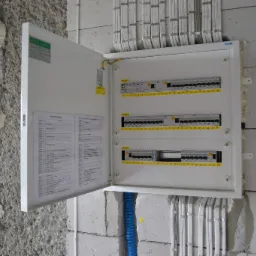 Otwarta skrzynka rozdzielcza z listwą zaciskową i opisem obwodów, zamontowana na surowej ścianie z widocznymi przewodami w peszlach.