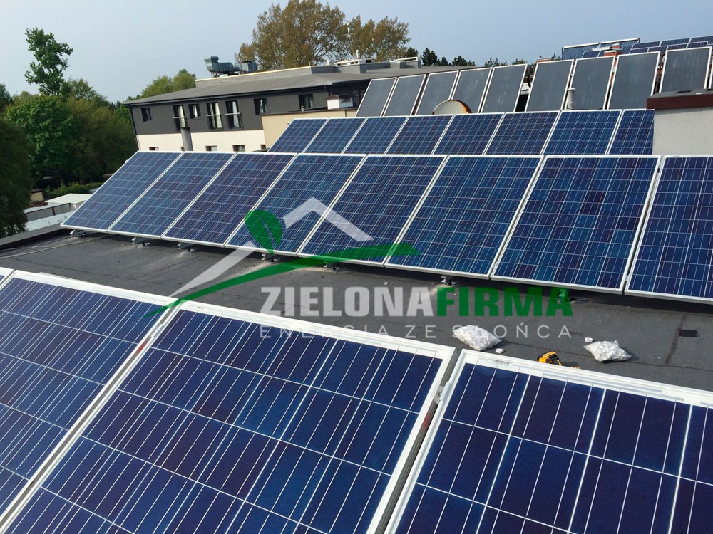 Panele słoneczne na dachu budynku, widoczne worki z elementami montażowymi i narzędzie, w tle inne panele słoneczne i budynek mieszkalny. Na panelach widoczne logo firmy.