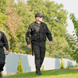 Dwóch ochroniarzy w czarnych uniformach patroluje teren zielony, jeden z krótkofalówką, drugi z pałką, na tle białego ogrodzenia i zieleni.