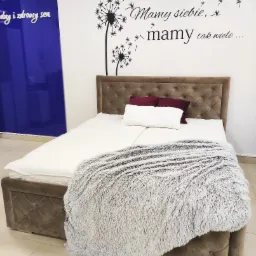 Tapicerowane łóżko z pojemnikiem, z pikowanym wezgłowiem i pluszowym kocem, na tle ściany z naklejką z dmuchawcami i napisem 'Mamy siebie, mamy tak wiele...'