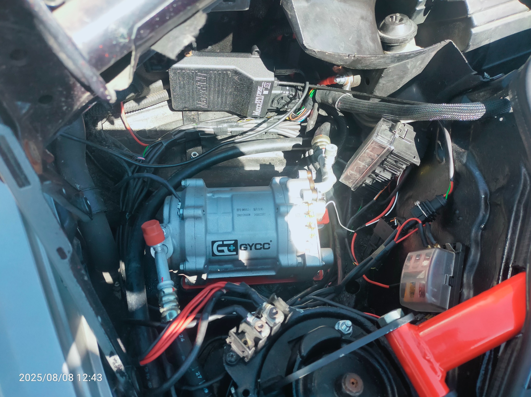 Silnik samochodowy z nową sprężarką klimatyzacji GYCC, widoczne przewody, bezpieczniki i inne komponenty. Ujęcie z góry, oświetlenie dzienne.