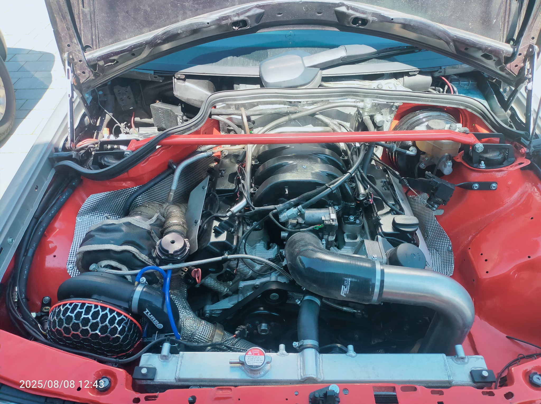 Otwarta maska czerwonego samochodu, widoczny silnik po tuningu z elementami chłodzenia i turbosprężarką. Czerwona rozpórka kielichów amortyzatorów.
