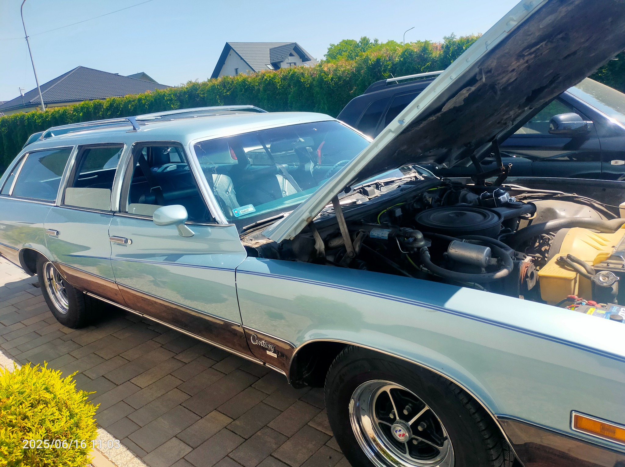 Błękitny, klasyczny kombi z otwartą maską prezentuje silnik. Widoczny akumulator i inne elementy. Tło: dom jednorodzinny i krzewy. Ujęcie z zewnątrz, w słoneczny dzień.