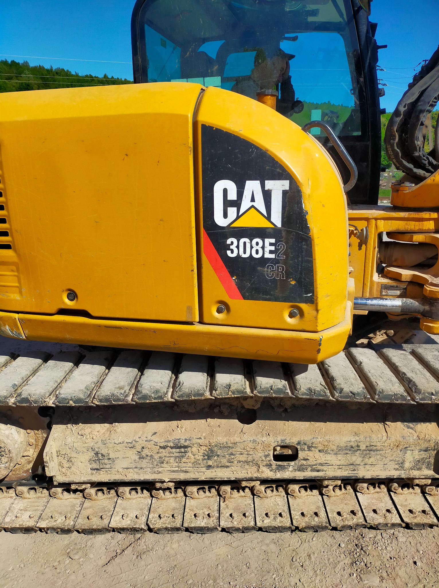 Żółta obudowa silnika koparki CAT 308E2 CR z widocznymi śladami błota na gąsienicach i obudowie, na tle błękitnego nieba i zieleni.