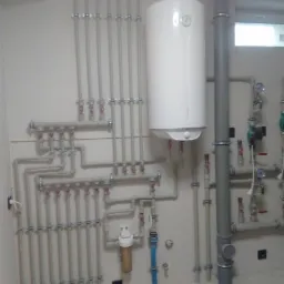 Biały bojler elektryczny Atlantic, liczne rury i zawory zamontowane na ścianie w pomieszczeniu technicznym, widoczny filtr wody i fragment okna.