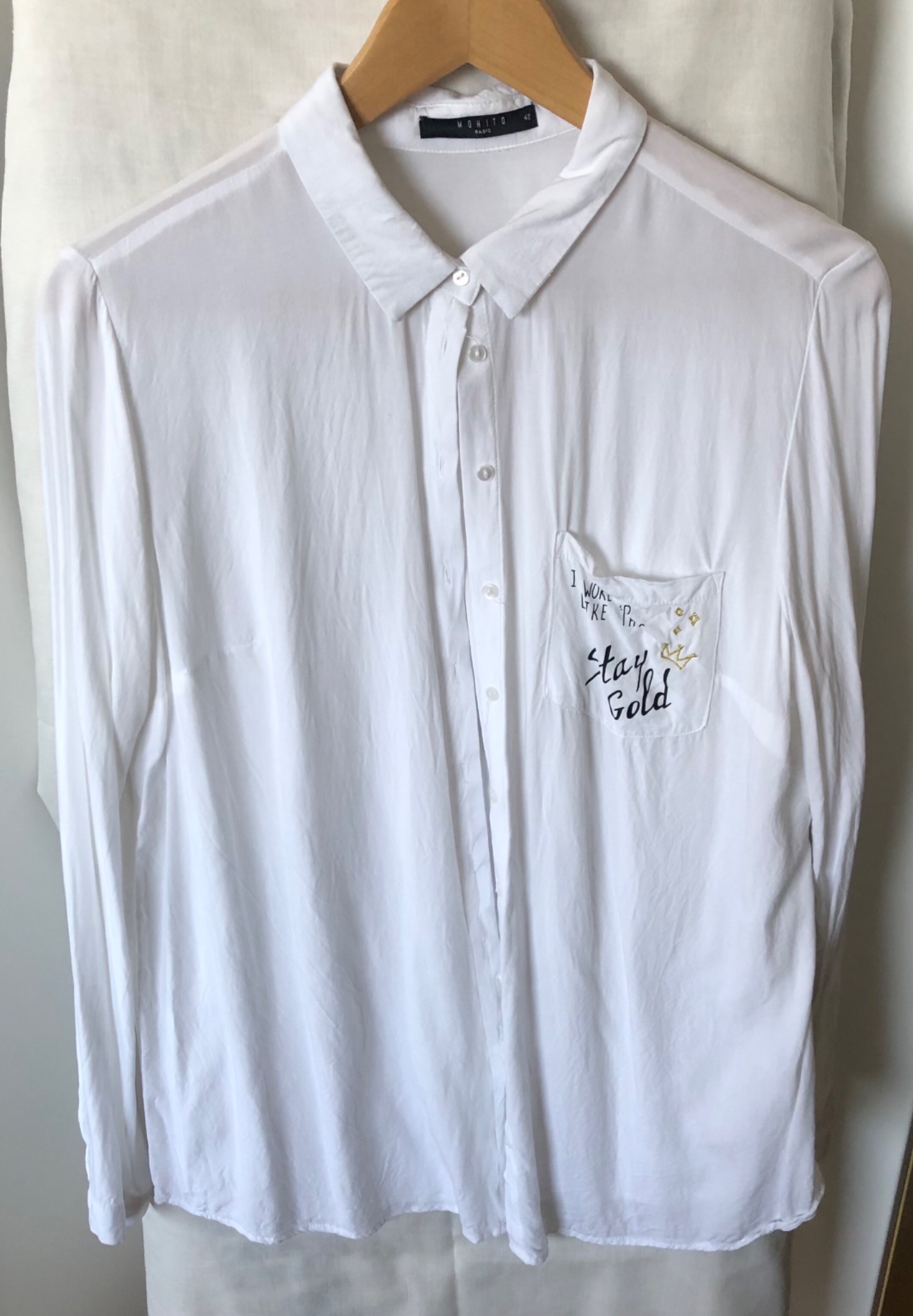 Biała, damska koszula z długim rękawem, wisząca na drewnianym wieszaku, z napisem 'Stay Gold' i motywem korony na kieszeni.