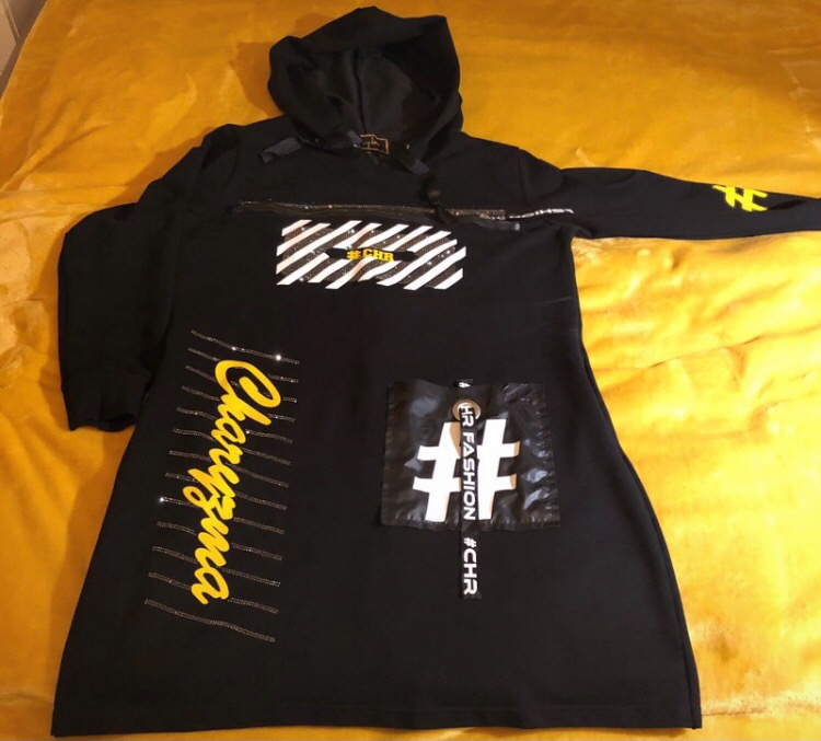 Czarna sukienka dresowa z kapturem, z aplikacjami i napisami w kolorze żółtym i białym, leżąca na żółtej narzucie.