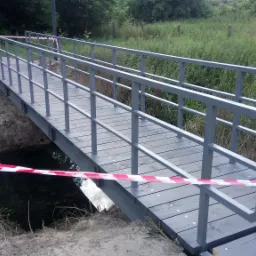 Szary, metalowy mostek nad ciekiem wodnym, zabezpieczony taśmą ostrzegawczą w kolorze biało-czerwonym, otoczony zielenią.