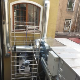 GARIN Sp. z o.o. - Zewnętrzna instalacja wentylacyjna z metalowymi rurami i centralą na dachu, widoczna drabina i okno budynku z żółtą elewacją.