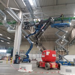 GARIN Sp. z o.o. - Montaż systemu wentylacyjnego w hali przemysłowej, pracownicy na podnośnikach koszowych instalują rury wentylacyjne pod sufitem.