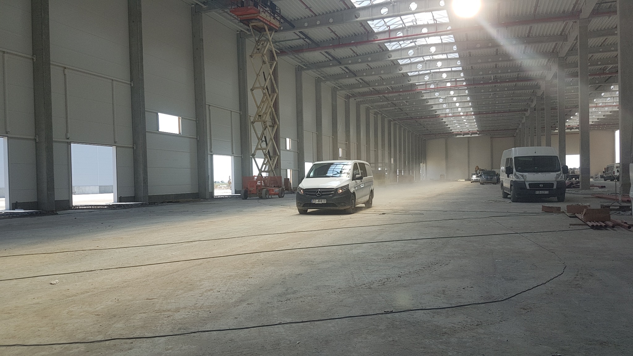 Wnętrze nowo budowanej hali magazynowej z widocznymi elementami wentylacji pod sufitem, betonową posadzką, pojazdami dostawczymi i podnośnikiem koszowym.