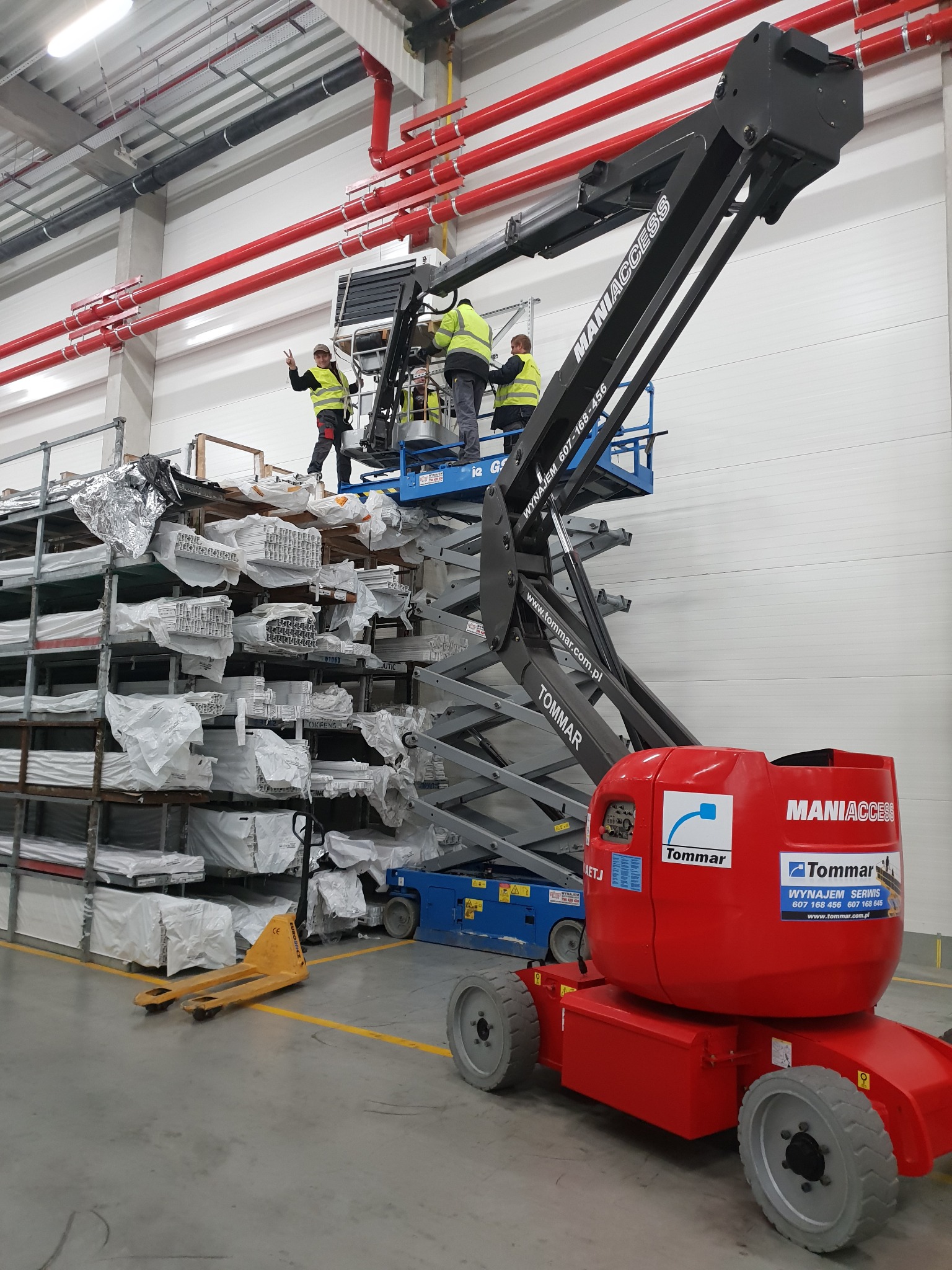 Montaż instalacji w magazynie: trzech pracowników na podnośniku koszowym MANIACCESS, instalacja rur pod sufitem, regały z materiałami budowlanymi w tle, jeden pracownik pokazuje znak zwycięstwa.
