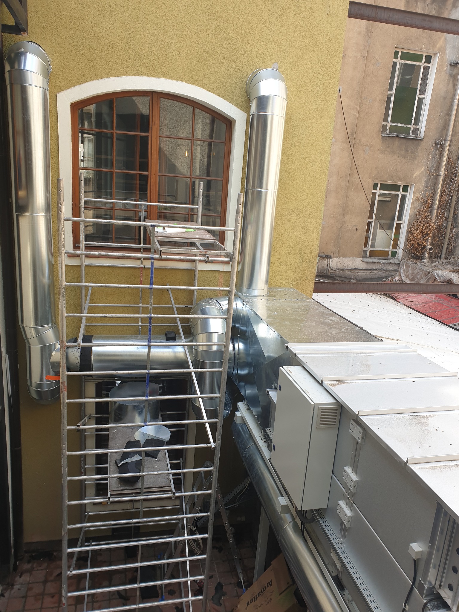 Zewnętrzna instalacja wentylacyjna z metalowymi rurami i centralą na dachu, widoczna drabina i okno budynku z żółtą elewacją.