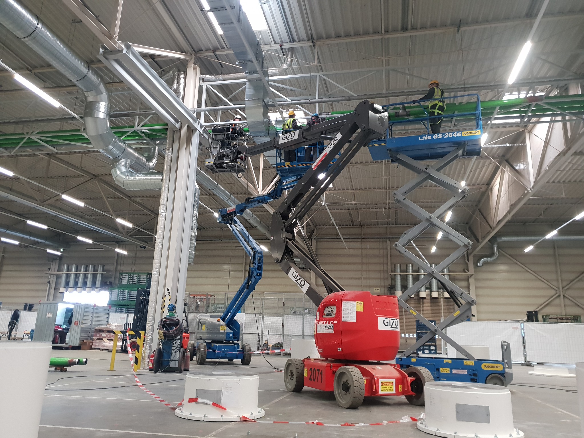 Montaż systemu wentylacyjnego w hali przemysłowej, pracownicy na podnośnikach koszowych instalują rury wentylacyjne pod sufitem.