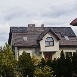 Dom jednorodzinny z brązowym dachem pokrytym panelami słonecznymi, widoczny przez gęste krzewy i drzewa iglaste.