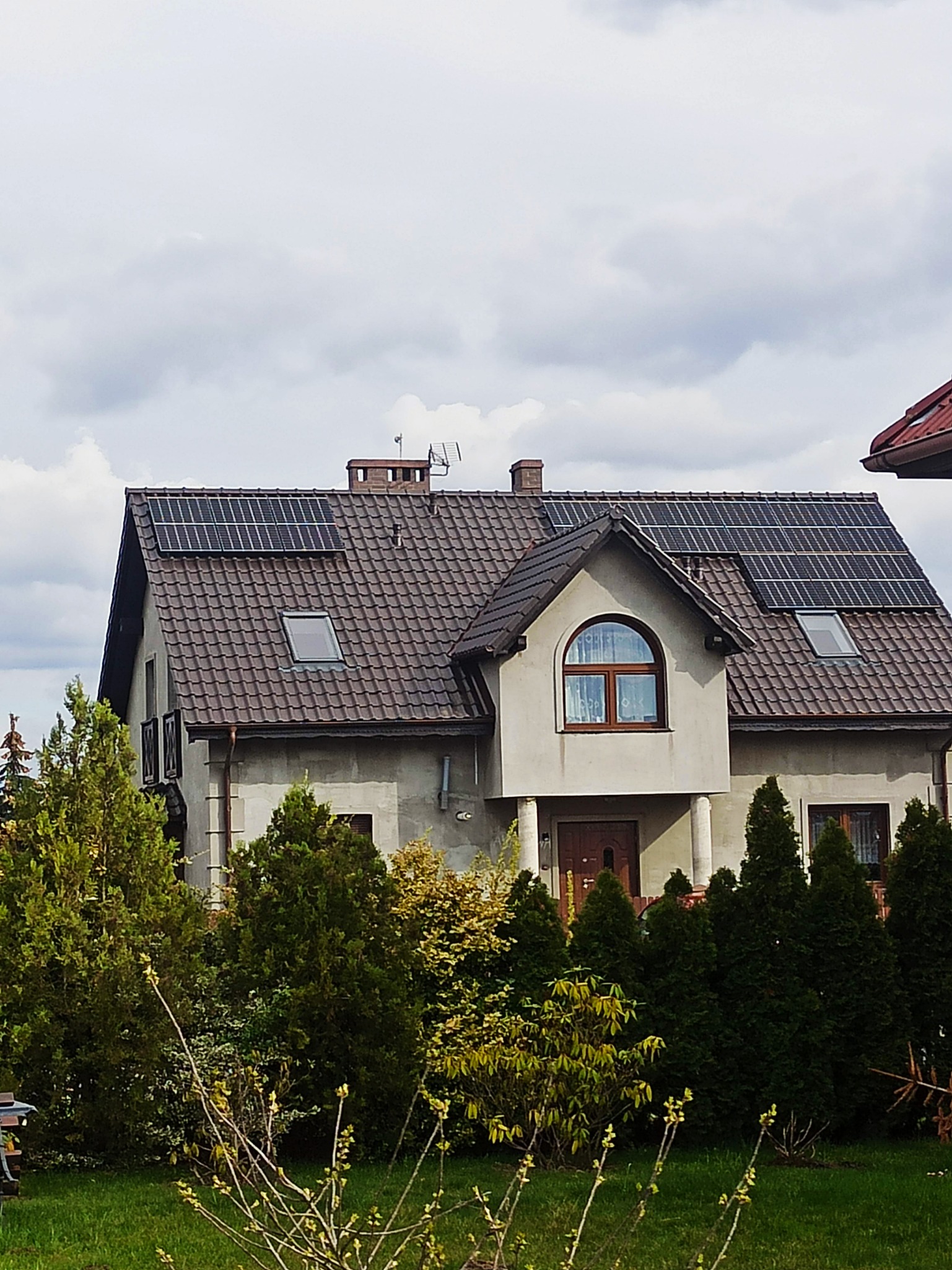 Dom jednorodzinny z brązowym dachem pokrytym panelami słonecznymi, widoczny przez gęste krzewy i drzewa iglaste.